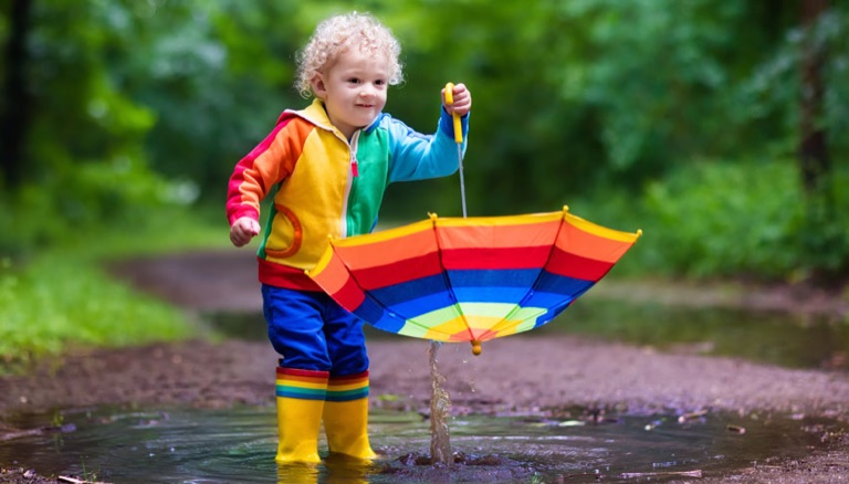 Kind in bunter Regenkleidung mit Schirm in einer Pfütze