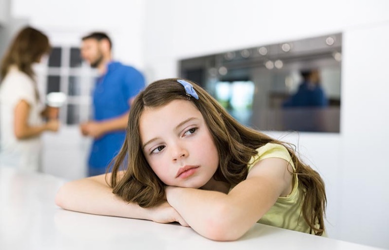 Mädchen sitzt niedergeschlagen am Tisch, die Eltern im Hintergrund