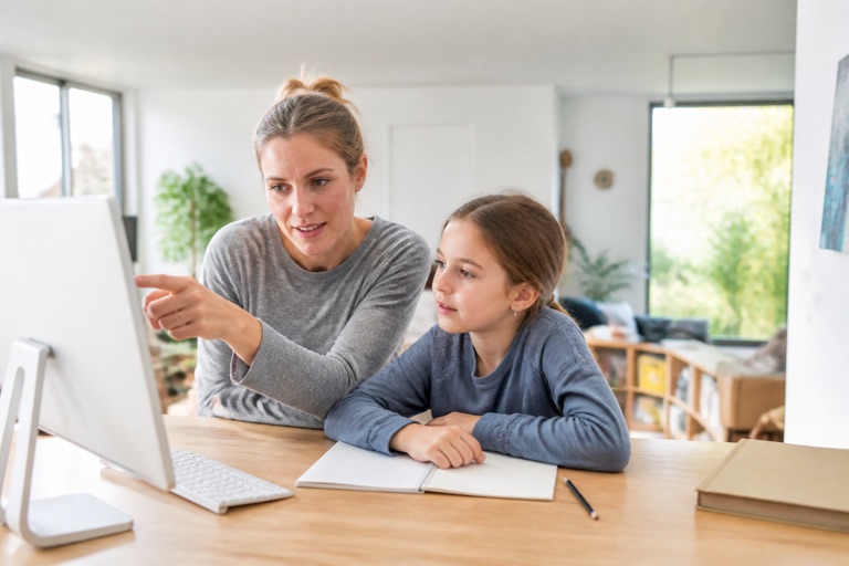 Elternteil sitzt mit einem Grundschulkind am Computer und erklärt etwas
