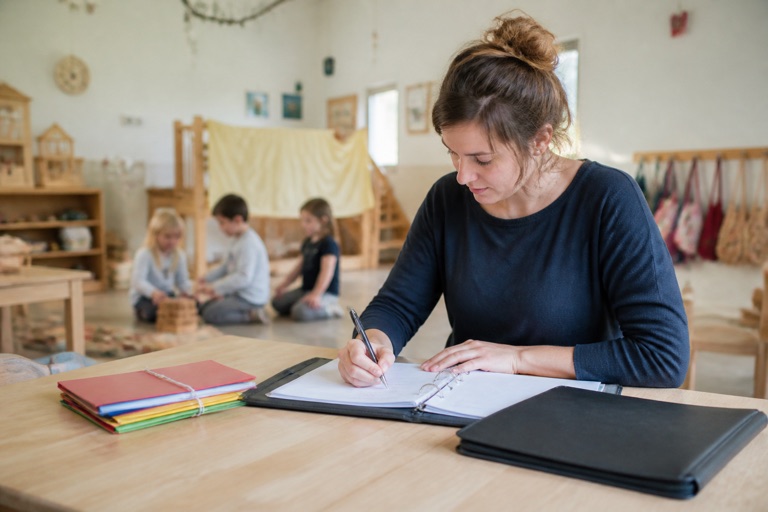 Erzieherin macht Notizen in einem Ordner, im Hintergrund spielende Kinder