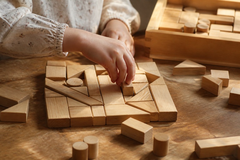 Hölzerne Fröbel-Spielgaben auf einem Tisch, eine Kinderhand legt einen Baustein